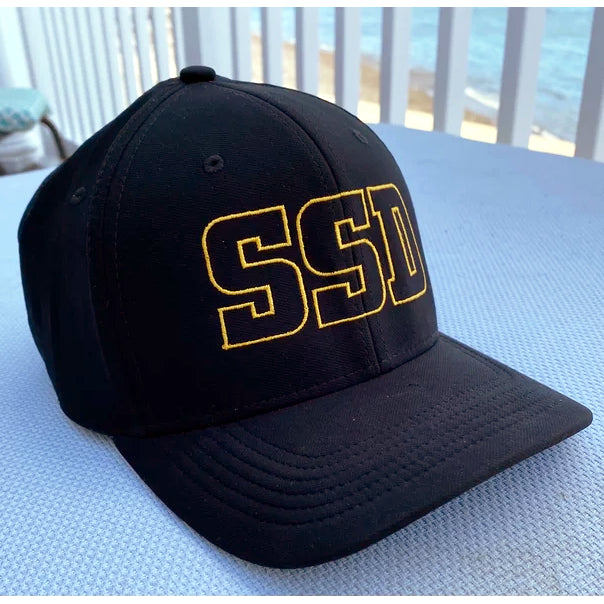 A black SS Decontrol baseball cap with large yellow outlined SSD letters and a Nike Swoosh is displayed on a light blue surface, set against a beach backdrop.