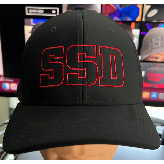 A black SS Decontrol AeroBill baseball cap with large red outlined SSD letters embroidered on the front. The background shows blurred images of other hats and colorful objects.