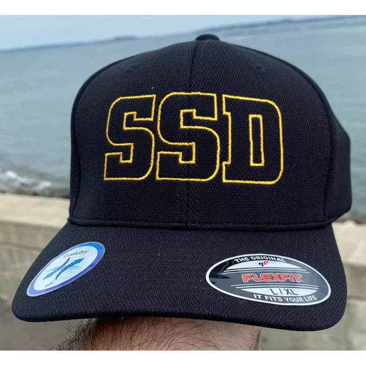 A black SS Decontrol baseball hat with bold yellow SSD gold embroidery on the front. The cap, sporting stickers on the brim, is displayed against a blurred outdoor background of water and sky.