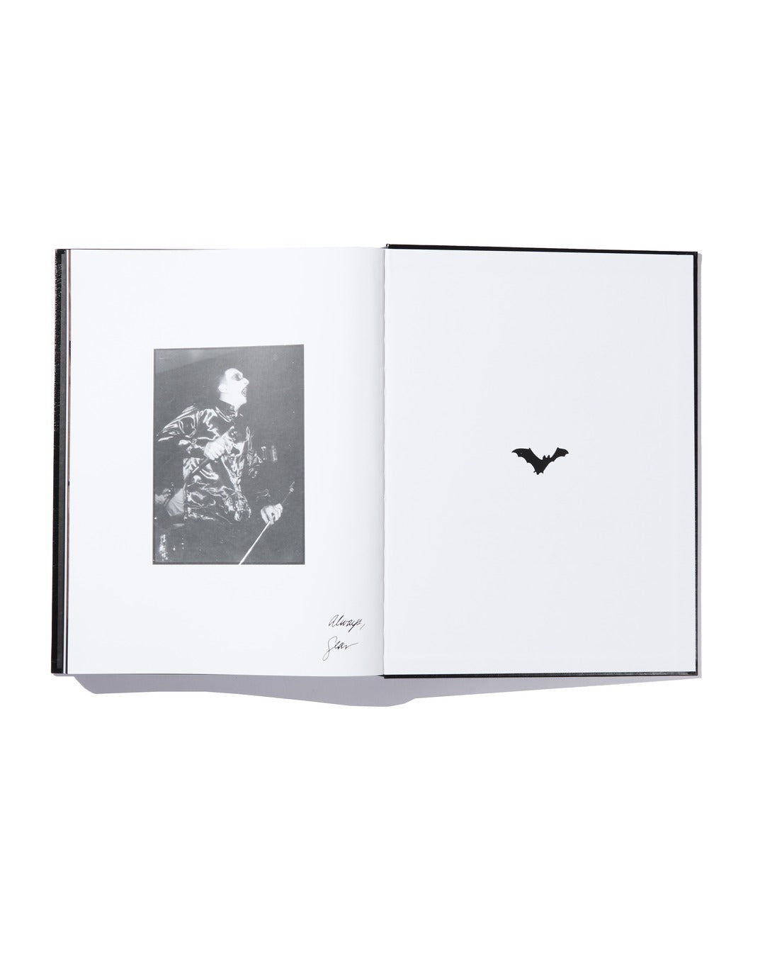 An open book from Ink & Dagger displays a black-and-white photo of a man singing on the left page with a signature underneath, while the right page features a small black bat illustration in the center. This limited edition hardcover highlights The Almighty Jeremy Dean Erik Bader.