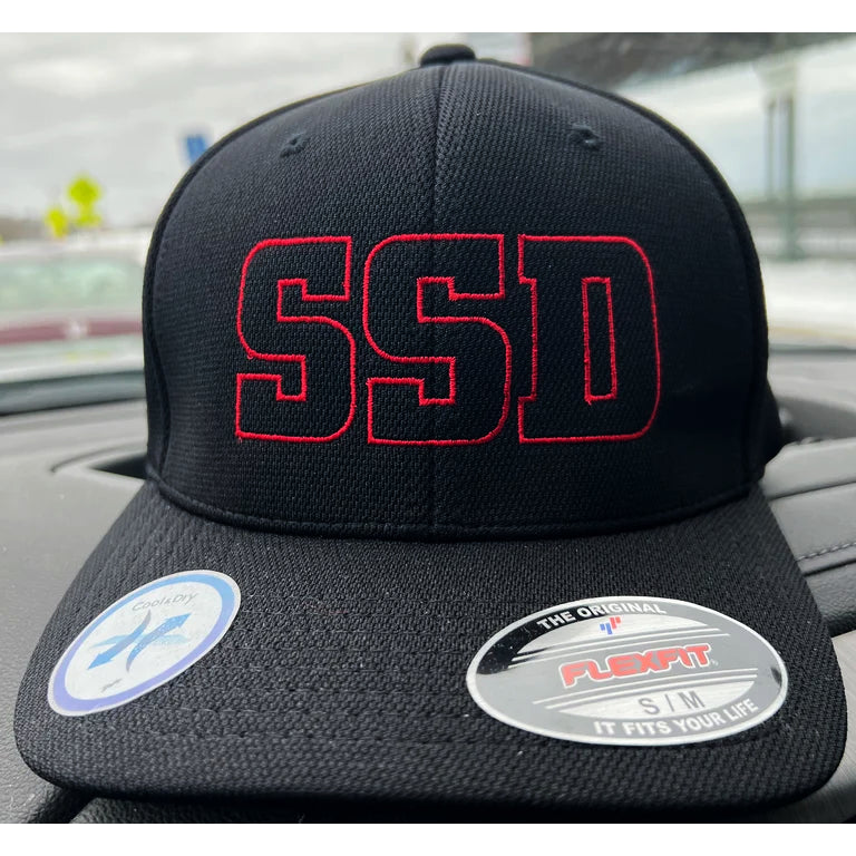 A black SS Decontrol Flexfit Baseball Hat with large red SSD embroidery and Cool & Dry Technology rests on a car dashboard, with stickers visible on the brim.