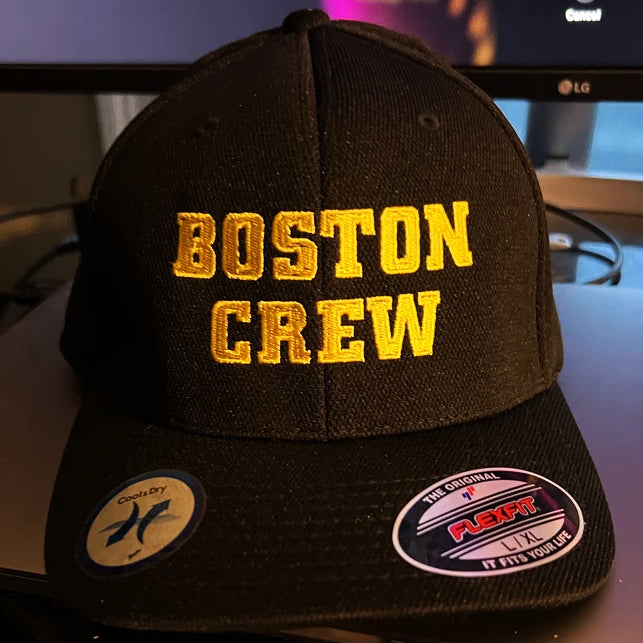 A black SS Decontrol Boston Crew hat with yellow embroidery, Cool &amp; Dry tech, and Flexfit LX stickers on the brim sits on a desk with a computer monitor in the background.
