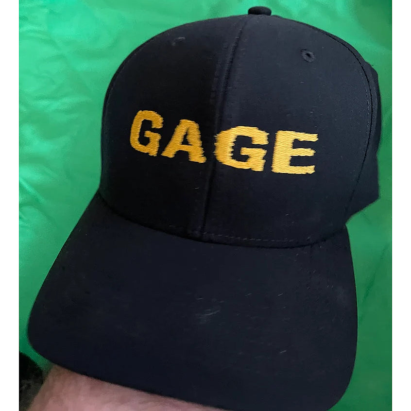 This lightweight summer hat by GAGE is black, features “GAGE” embroidered in yellow on the front, and is showcased against a green background.