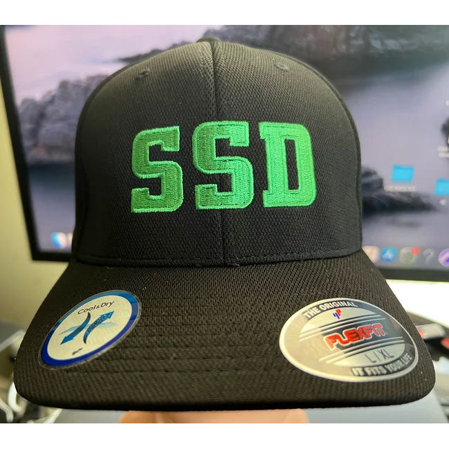 A SS Decontrol Flexfit Baseball Hat with bold green “SSD” embroidery on the front. Two stickers display the Flexfit logo and Cool & Dry Technology, with a blurred computer screen in the background for a modern vibe.