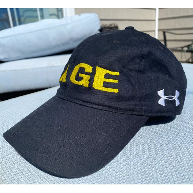 This black GAGE logo hat rests on a light blue cushion. The front features large yellow stitched letters “AGE.”.