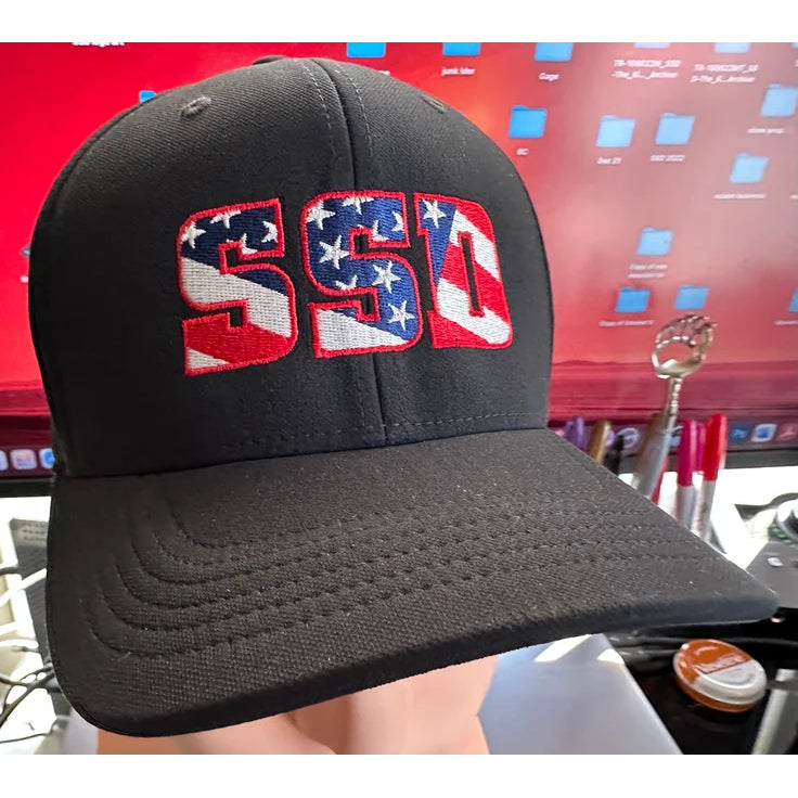A black SS Decontrol Classic 99 Hat with SSD embroidered in an American flag motif, shown before a red desktop screen, features Dri-FIT technology for added comfort.