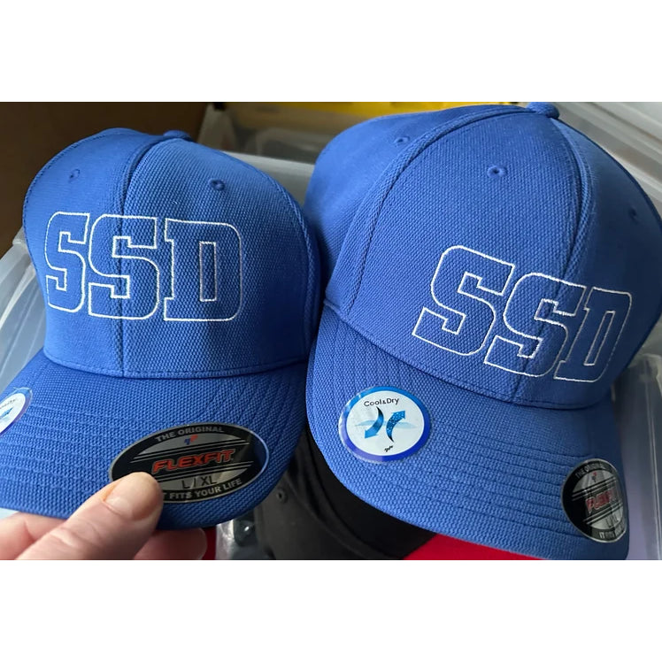 Two SS Decontrol blue baseball hats with SSD in white outline are displayed on a storage bin: one has a Flexfit Cool & Dry sticker, and the other features Poly Block Mesh.