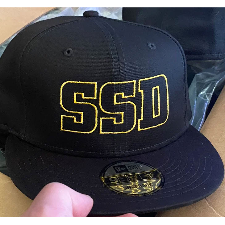 A black SS Decontrol snapback cap with a flat bill, bold yellow SSD embroidery on the front, and an adjustable fit. A shiny sticker accents the brim as a hand holds it.