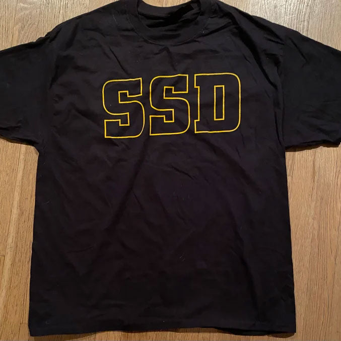 A black SS Decontrol Haynes Beefy T heavyweight t-shirt with large, bold yellow outlined SSD letters across the chest, laid flat on a wooden floor.