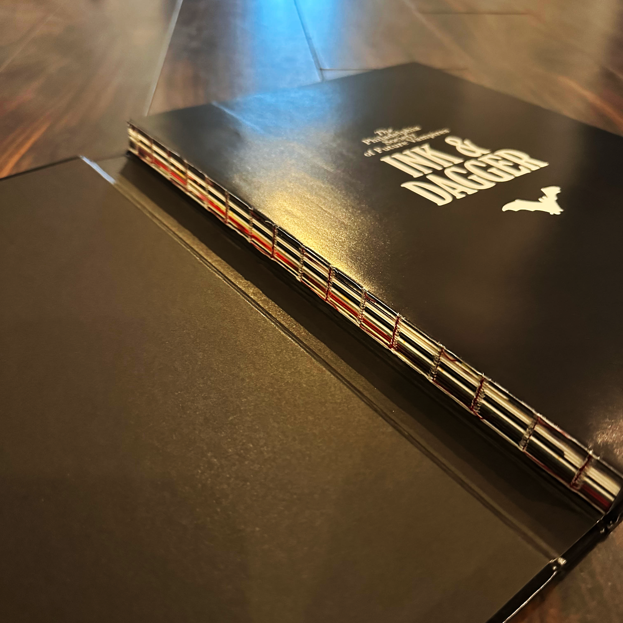 A close-up of an open Ink & Dagger Complete Works book displays its exposed multi-colored stitching along the spine as it rests on a wooden surface. This limited edition hardcover from Ink & Dagger exemplifies masterful craftsmanship and captivating design.