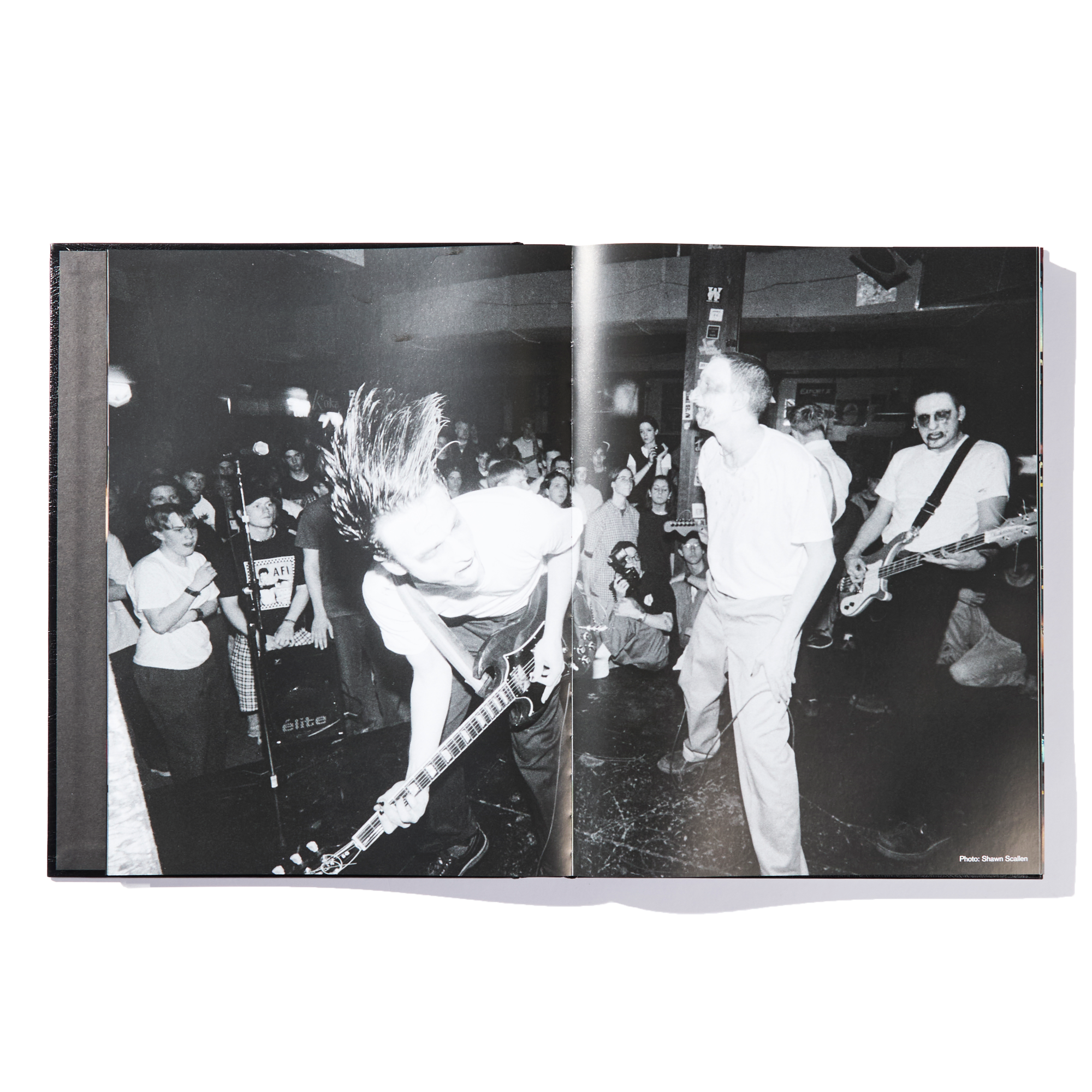 Black-and-white photo of a punk rock band performing on stage, with an energetic crowd in the background. One guitarist bends forward mid-strum as the singer and bassist engage with the audience, evoking the raw energy of an Ink & Dagger show—an ideal capture for any limited edition print or inclusion in The Almighty book from Ink & Dagger.