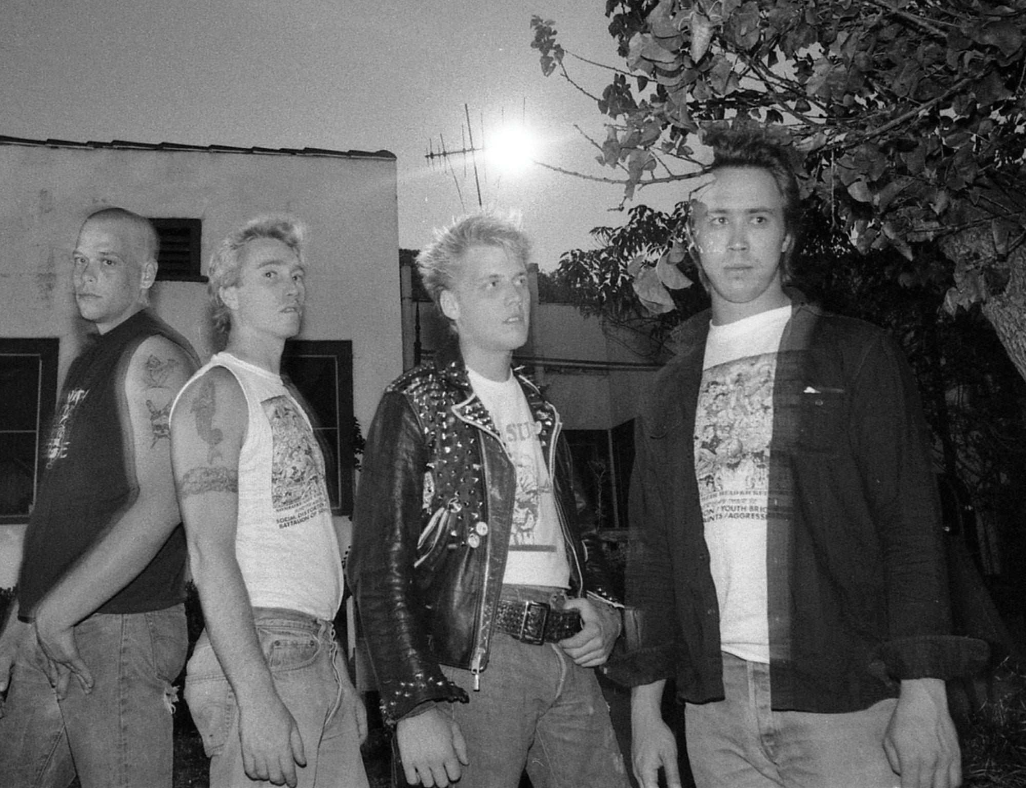 Four men stand outdoors at night—two with short blond hair in punk style, one in a studded leather jacket (Limited Edition) by Agression, the others in graphic t-shirts by the same brand—with a building and tree behind them.