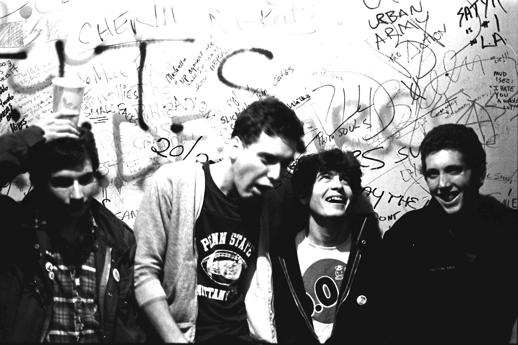 Four young men stand in front of a graffiti-covered wall; one holds a cup on his head, another leans forward with his mouth open, and the group appears to be laughing and having fun. The image is black and white.