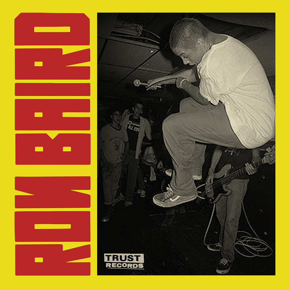 A black-and-white photo of a man jumping mid-air with a microphone at a live music show. Band members and audience are visible. Bold red text reads RON BAIRO on a yellow border. TRUST RECORDS logo appears at the bottom.