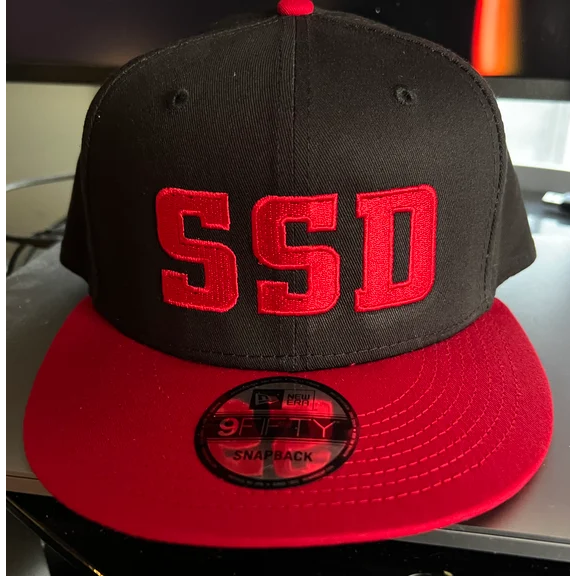 A black SS Decontrol New Era 9Fifty SnapBack hat with a bright red flat brim features the Red Solid SSD Logo embroidered on the front. The snapback sticker is visible on the brim as it sits on a dark surface.