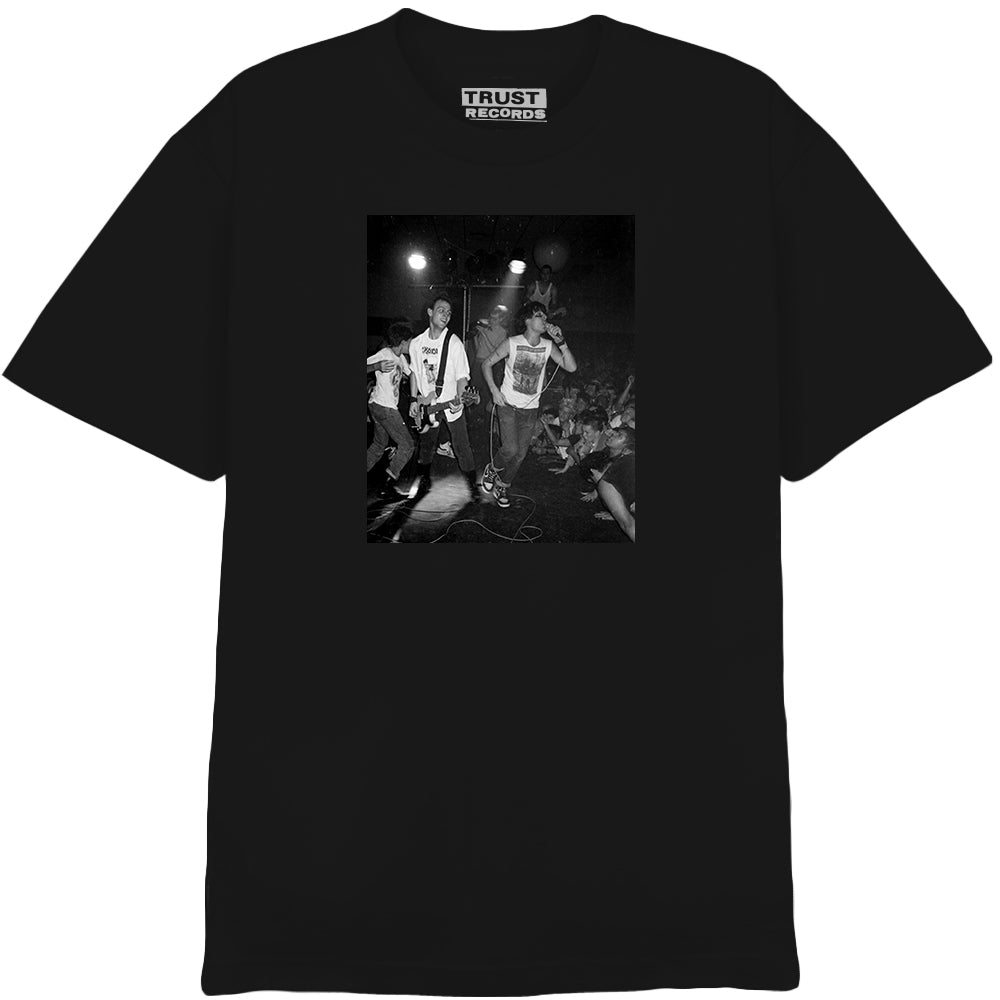 Black T-shirt from 7 Seconds featuring a silkscreened black and white live punk band photo with energetic members and a cheering crowd. Trust Records tag. Printed on Alstyle 1301 for premium quality.