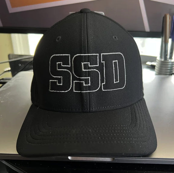 A black SS Decontrol Summer Hat with large, white, outlined SSD letters embroidered on the front. This comfy AeroBill cap sits on a metallic surface, electronic gear in the background.