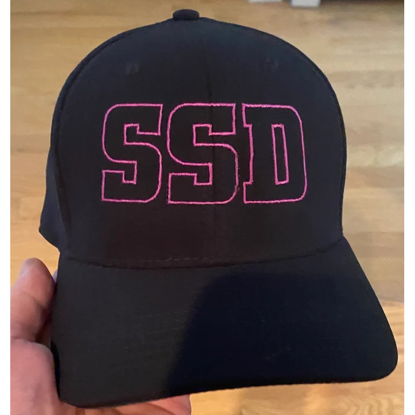 A black SS Decontrol Dri-FIT hat with SSD outlined in pink on the front is held up by a hand, with a wooden floor in the background.