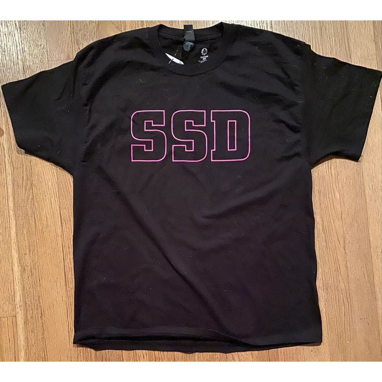 A black SS Decontrol tee is laid flat on a wooden floor, displaying large SSD lettering in purple outline centered on the chest.