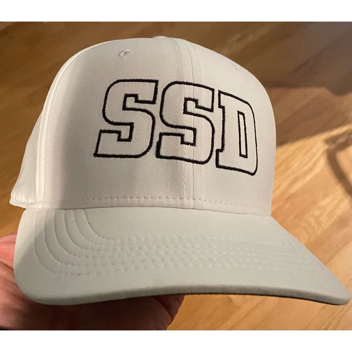A SS Decontrol Classic 99 white baseball cap by Nike, with bold black SSDecontrol letters embroidered on the front, AeroBill and Dri-FIT tech, held by hand over a wooden floor.