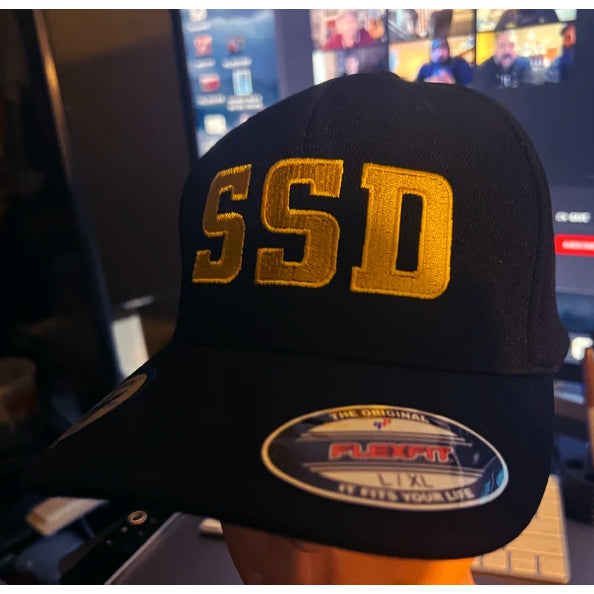 A black SS Decontrol Flexfit baseball hat with large SSD letters in yellow gold embroidery sits on a desk, with a computer monitor showing a blurred video call in the background.