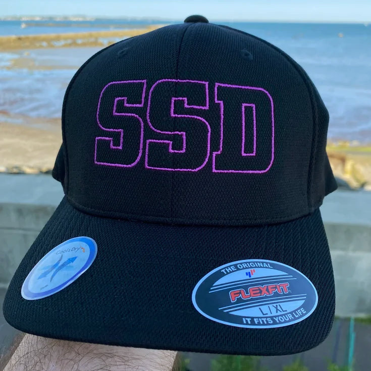 A black SS Decontrol Flexfit Cool & Dry baseball hat featuring large purple SSD letters on the front, shown close-up. FLEXFIT and sizing stickers are visible on the brim, with a beach and water in the background.