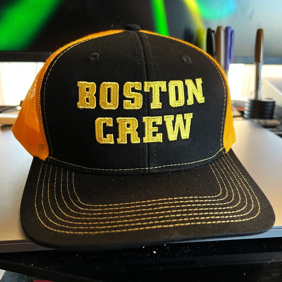A SS Decontrol Black and Gold Trucker Cap with BOSTON CREW embroidered in yellow rests on a desk with pens and a monitor behind it. This adjustable mesh cap brings urban flair to any workspace.