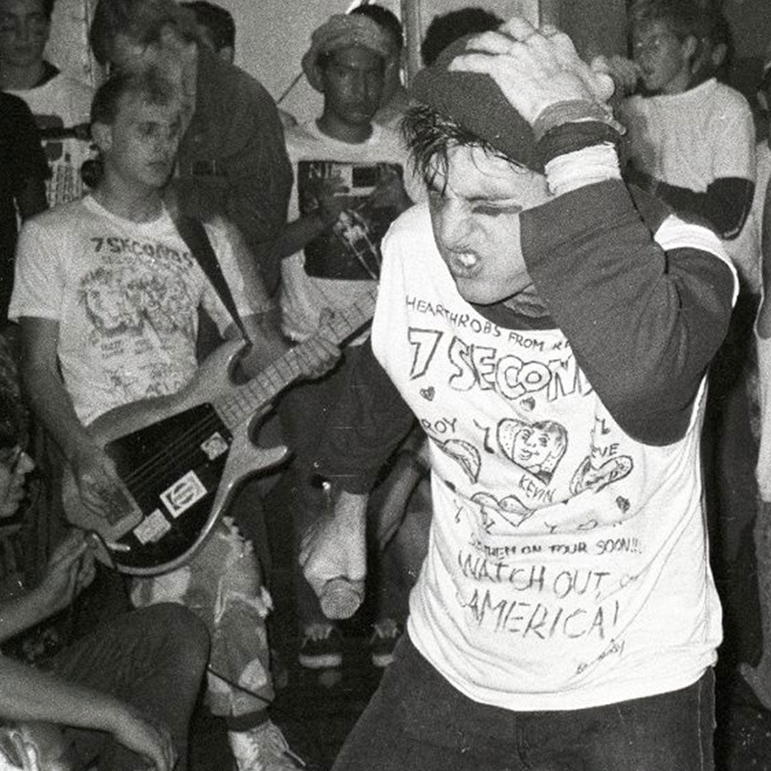 Black and white photo of a punk band playing live. The vocalist sings passionately as a bassist in a Brian Walsby tee plays nearby. Both wear 7 Seconds shirts, representing the 7 Seconds brand while performing for an engaged crowd.