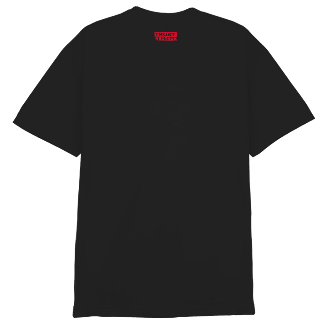 A plain black 100% cotton T-shirt by 7 Seconds, viewed from the back, features a small red silkscreened label with white "TRUST NOBODY" text near the neck.