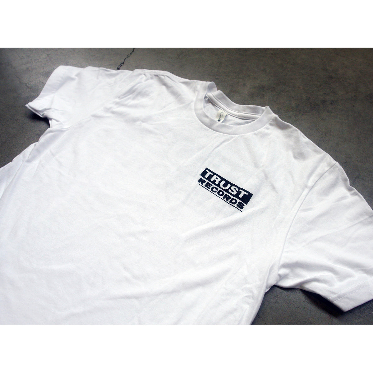 A White Trust Records T-Shirt rests on a gray surface, showcasing a small black rectangular logo patch with white text on the left chest.