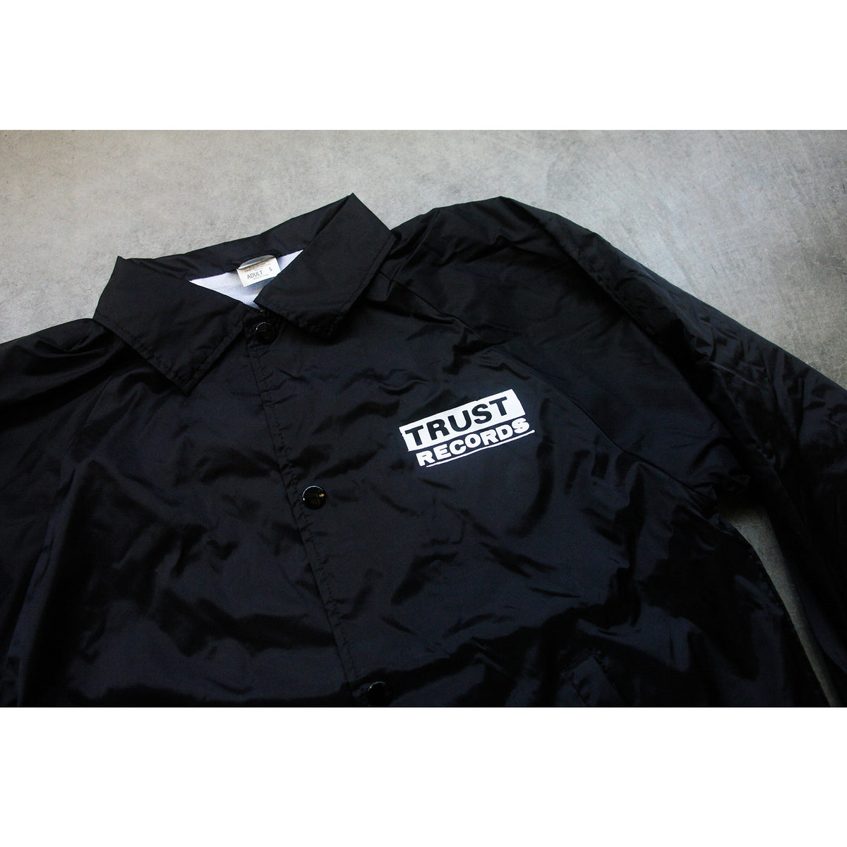A black Trust Records windbreaker with a collar and a white chest patch reading TRUST RECORDS in bold black text is laid flat on concrete.