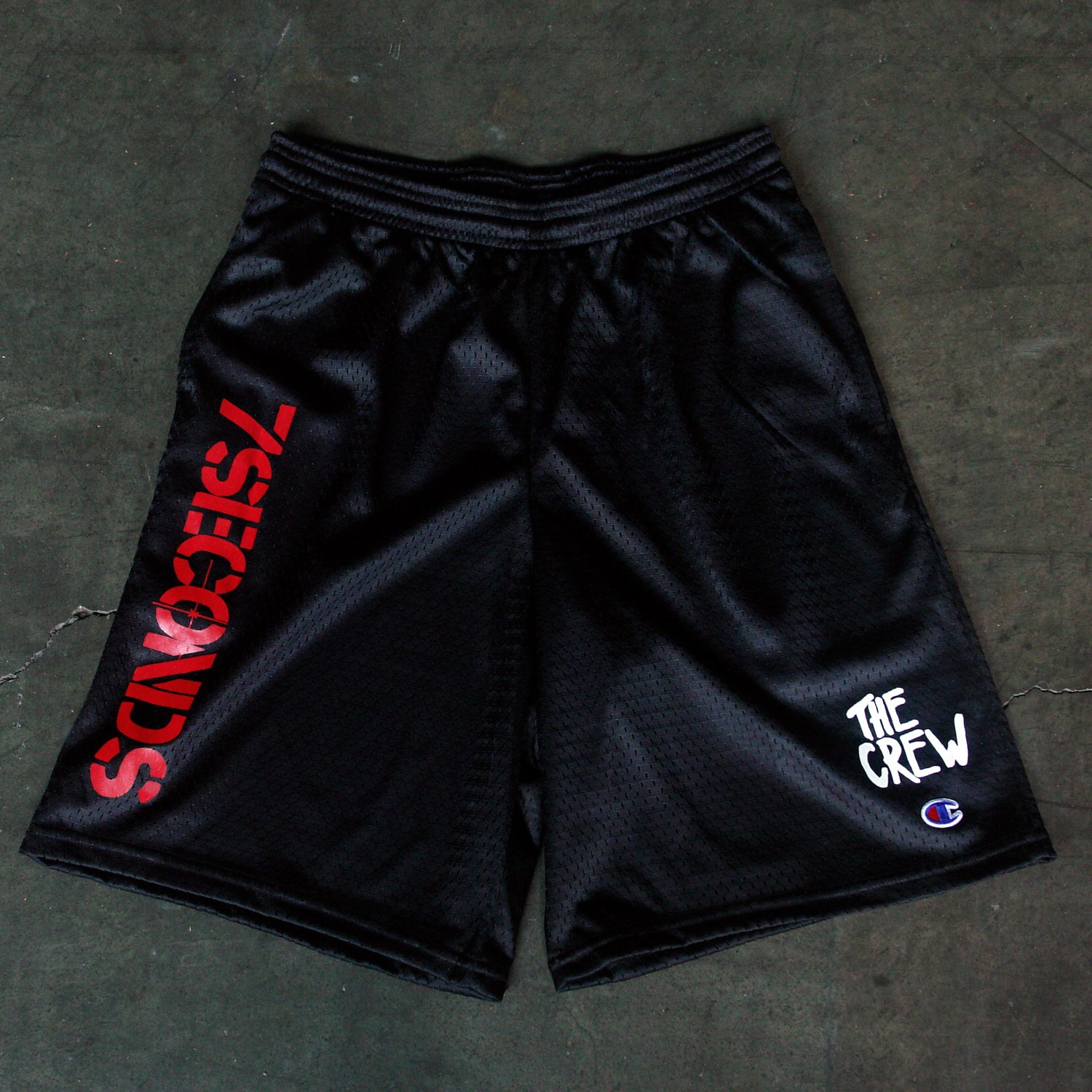 Black mesh Champion shorts by 7 Seconds with 7SECONDS in bold red on the left leg, THE CREW in white graffiti font on the right, plus side pockets and a Champion logo near the right hem.
