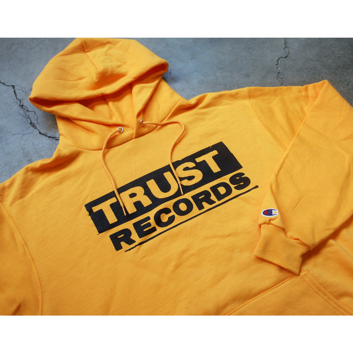 A yellow Champion Pullover hoodie by Trust Records with "TRUST RECORDS" in bold black letters on the front, shown on a textured gray surface. The left sleeve features the signature Champion logo.