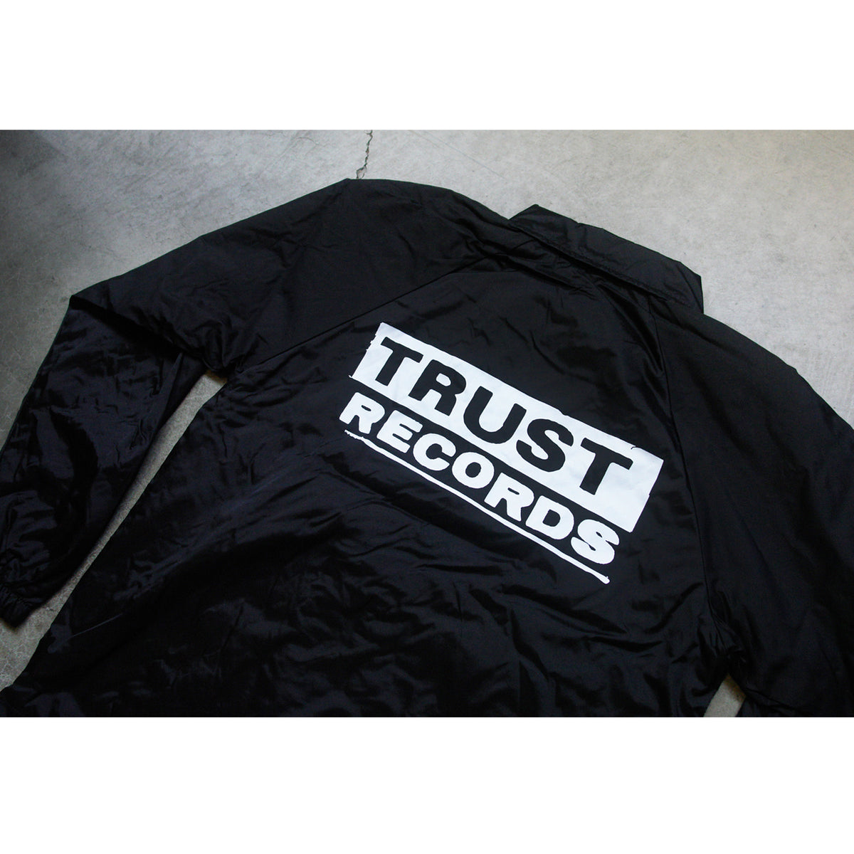 A black Trust Records windbreaker is laid flat on concrete, with TRUST RECORDS boldly printed in white across the back.