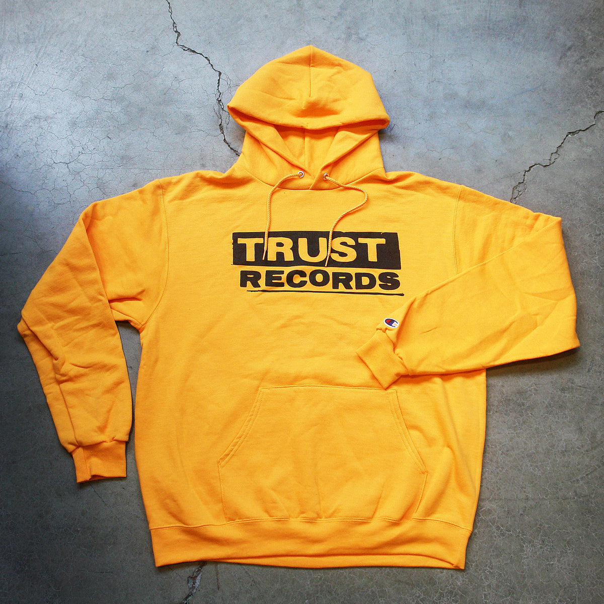 A bright yellow Trust Records pullover hoodie with TRUST RECORDS in bold black letters on the front, laid flat on a gray concrete surface.