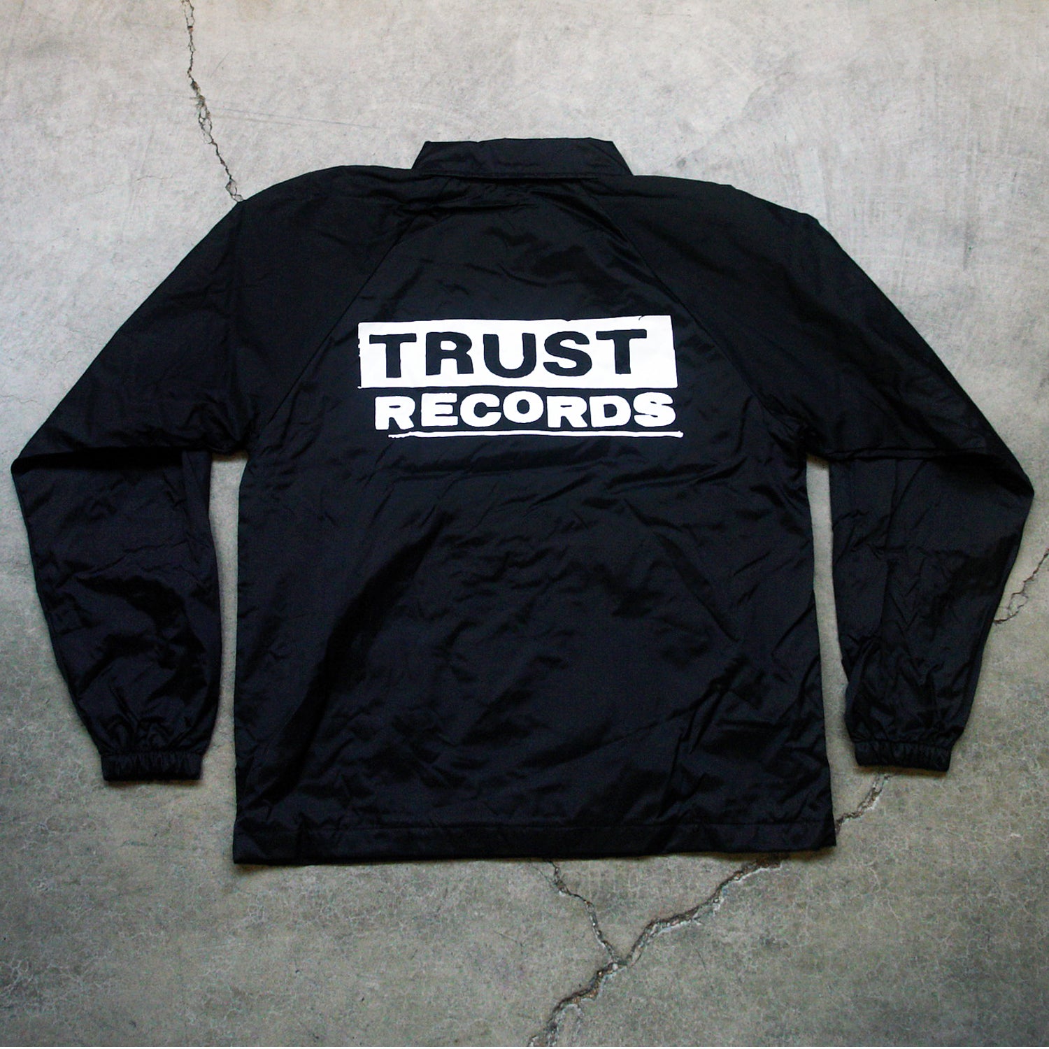 A black windbreaker from Trust Records features TRUST RECORDS printed in bold white letters on the back and is displayed flat on a cracked concrete surface.