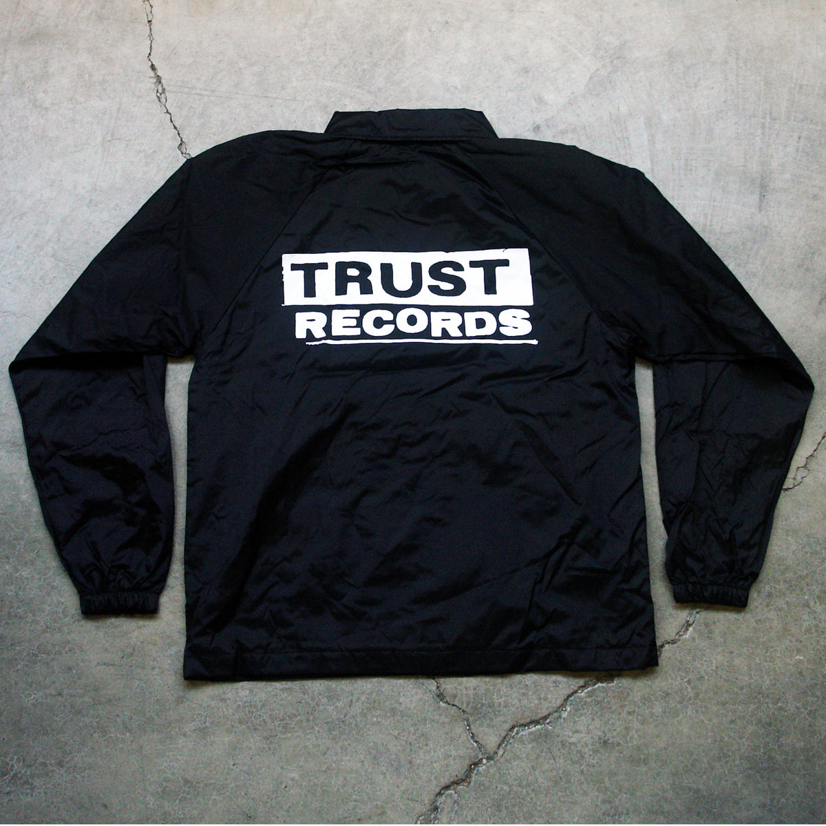 A black windbreaker from Trust Records features TRUST RECORDS printed in bold white letters on the back and is displayed flat on a cracked concrete surface.
