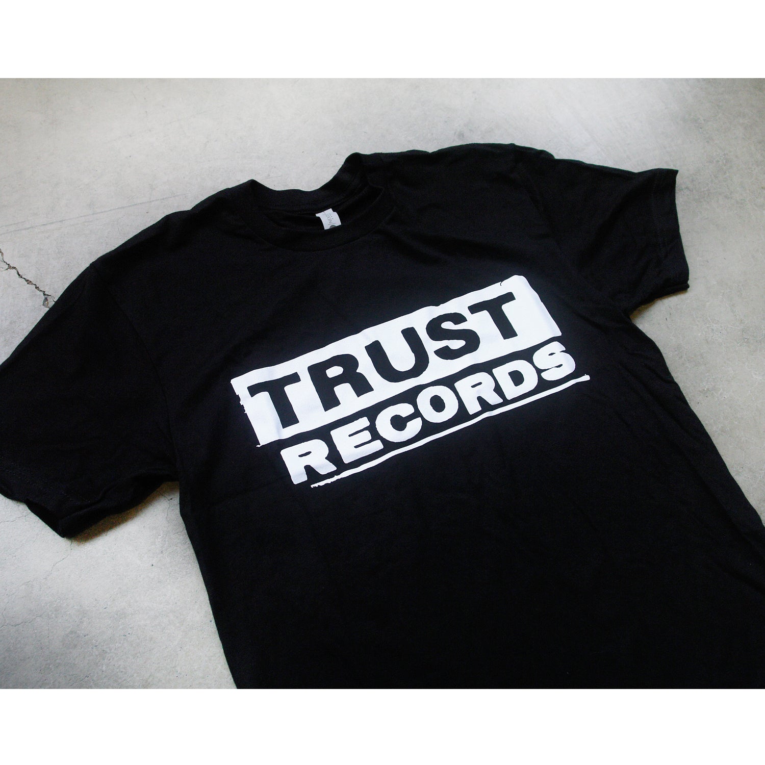 A black Trust Records T-shirt lays flat on a light surface, showcasing a bold white “TRUST RECORDS” logo in all caps across the chest.