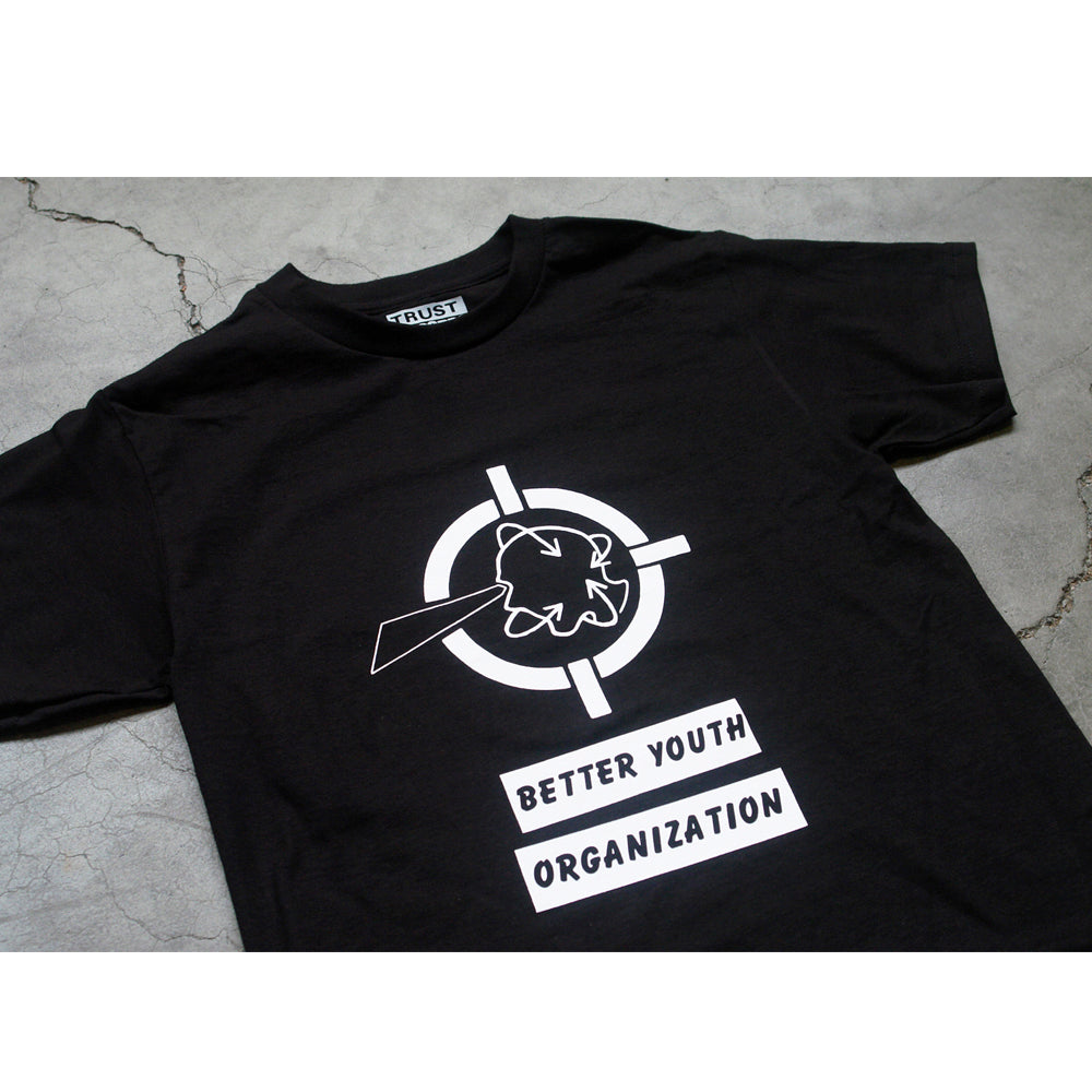 Black t-shirt by Better Youth Organization laid flat on a gray surface, featuring a silkscreened white graphic of a flower with a diagonal line inside a crosshair and “BETTER YOUTH ORGANIZATION” text below the design.
