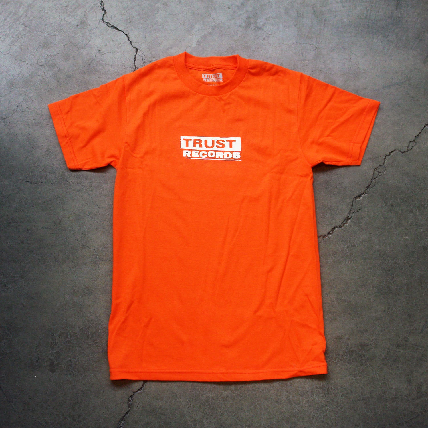 An orange Trust Records Alstyle 1301 T-shirt with “TRUST RECORDS” in bold white letters inside a white rectangle, silkscreened on 100% cotton for comfort and durability, laid flat on cracked concrete.