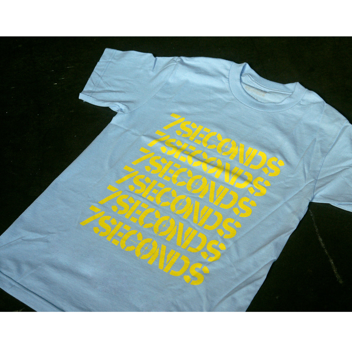 A light blue 100% cotton T-shirt from 7 Seconds, featuring 7 SECONDS printed repeatedly in bold yellow letters diagonally across the front, is silkscreened and shown laid flat on a dark surface.