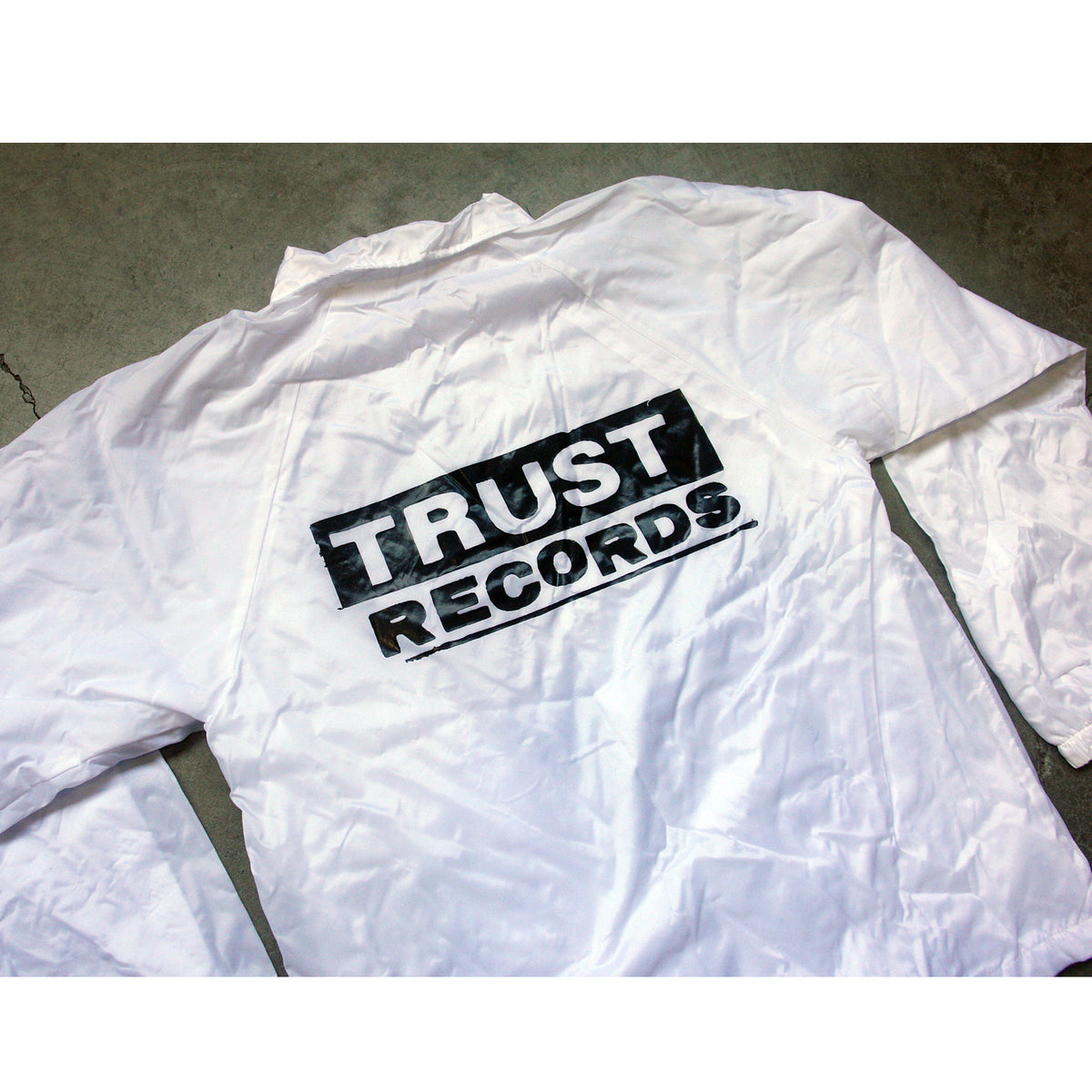 A white windbreaker from Trust Records rests on a concrete surface, featuring the brand's name printed boldly in black across the back.