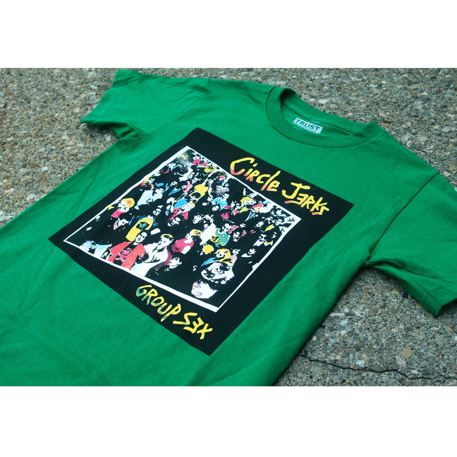 A green Circle Jerks t-shirt featuring colorful cartoon crowd art, silkscreened with “Circle Jerks” at the top and “Group Sex” at the bottom, laid flat on a textured outdoor surface.