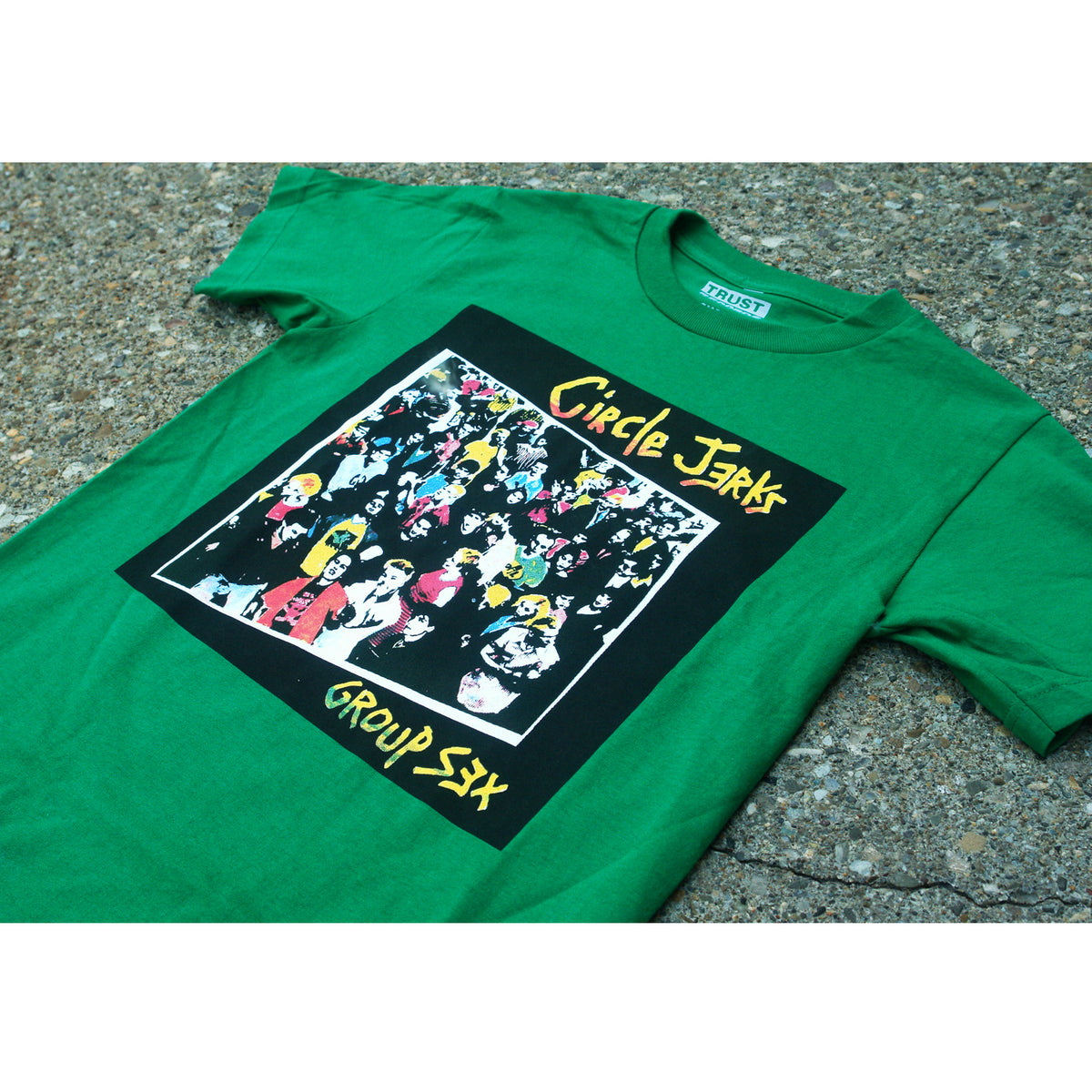 A green Circle Jerks t-shirt featuring colorful cartoon crowd art, silkscreened with “Circle Jerks” at the top and “Group Sex” at the bottom, laid flat on a textured outdoor surface.
