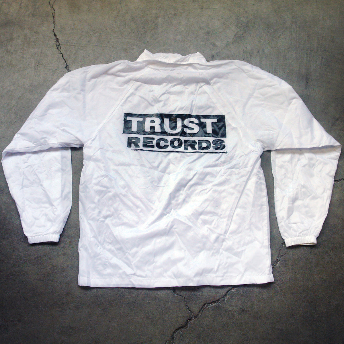 A white, long-sleeve windbreaker from Trust Records lies flat on concrete, with a bold black-and-white TRUST RECORDS logo printed in large capital letters across the back.