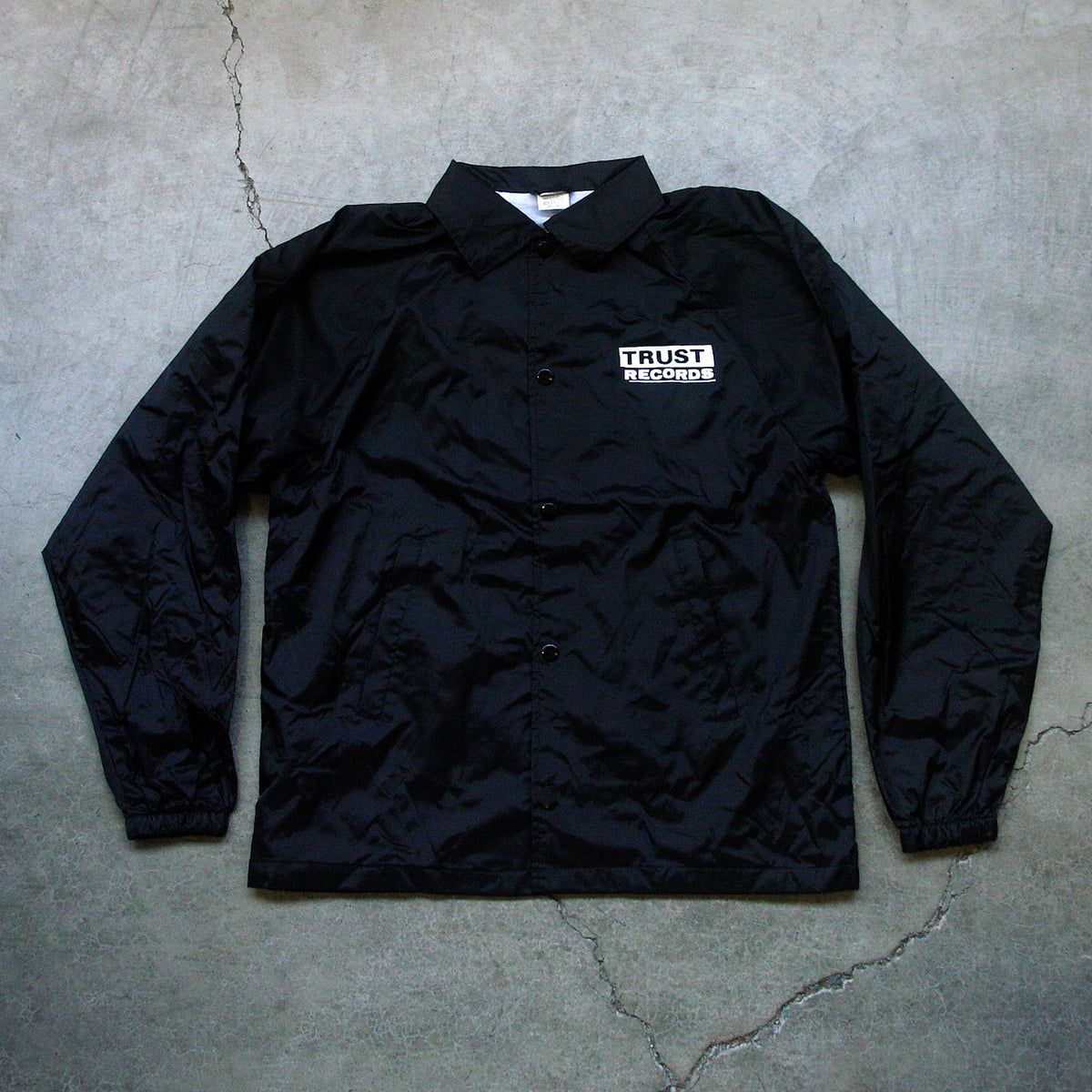A black Trust Records windbreaker with long sleeves, featuring a white logo patch on the left chest, displayed on concrete.