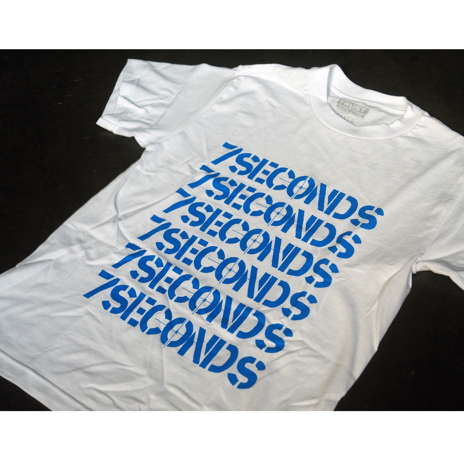 A white 7 Seconds Alstyle 1301 T-shirt, made of 100% cotton, features 7SECONDS repeatedly silkscreened in large blue capitals diagonally on the front.
