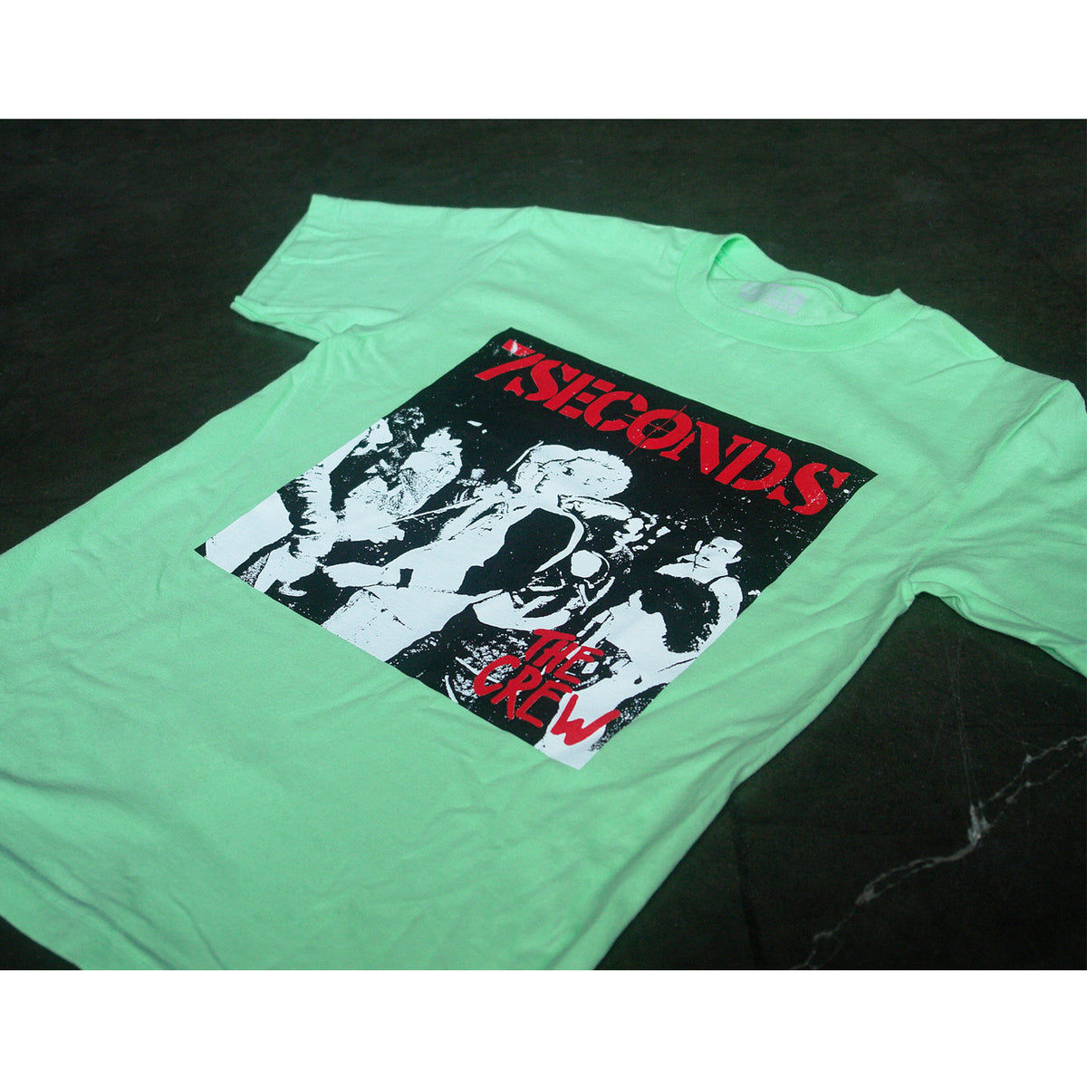 A light green 7 Seconds Alstyle 1301 T-shirt, featuring a black-and-white punk concert crowd graphic and red "7SECONDS" and "THE CREW" text on the front, is laid flat on a dark surface.