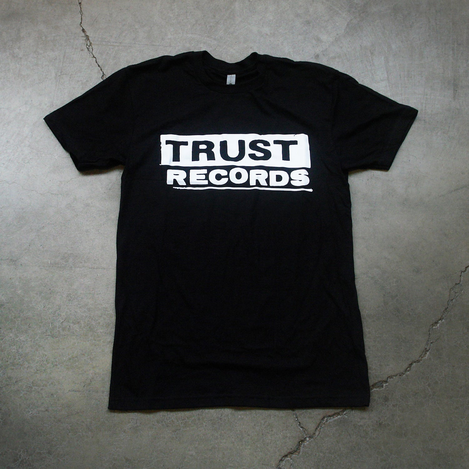 A Trust Records Black T-Shirt features the White Logo printed inside a rectangular box, displayed flat on a gray concrete surface.