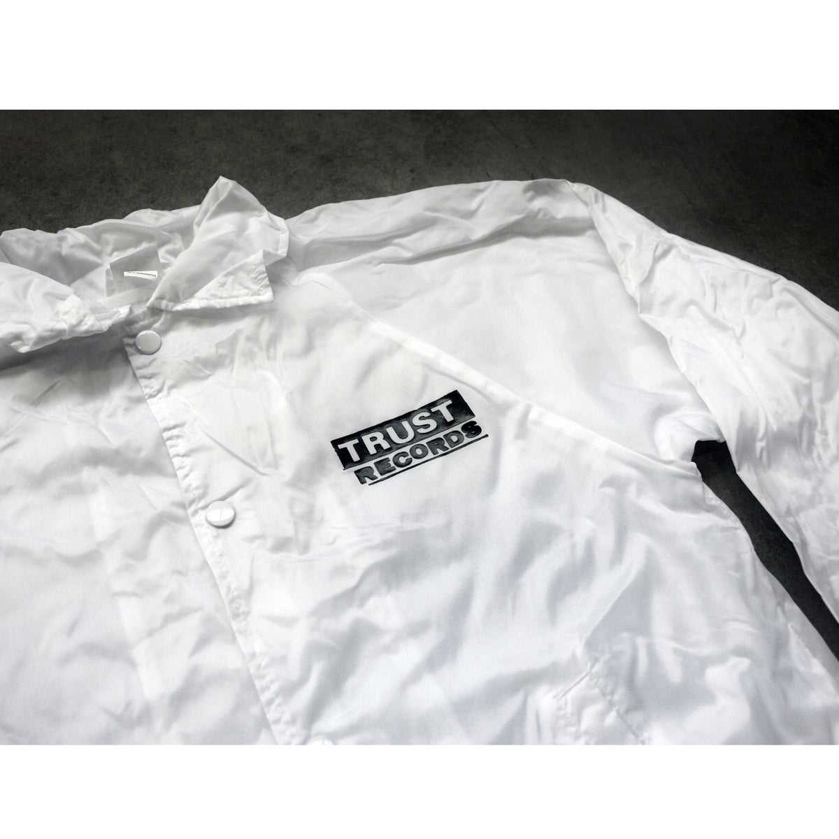 A white Trust Records windbreaker jacket rests on a gray surface, featuring a black and white chest patch that reads TRUST RECORDS.